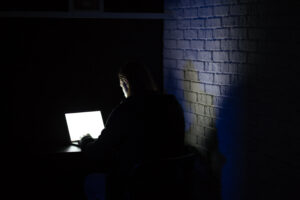 In a black and white setting, a young man is seated in a dimly lit room, searching for a computer. The man appears suspicious, his face partially obscured in shadow, creating a mysterious and secretive ambiance.